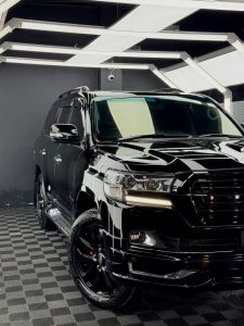 A glossy black Toyota Land Cruiser 200 Series SUV parked indoors on a patterned tile floor, showing the front three-quarter view with bright overhead lighting.